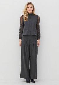 Woman wearing black high-neck blouse with lace sleeves and wide-leg black pants, standing against a plain white wall.