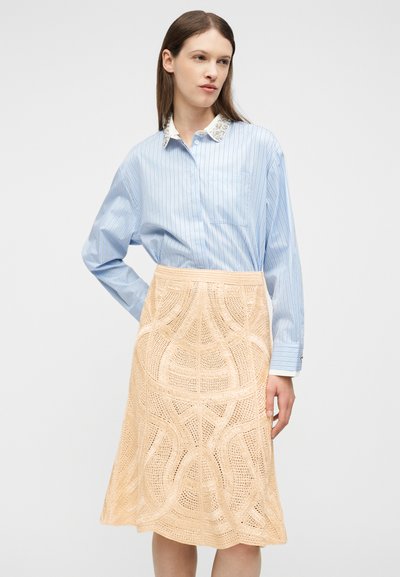 Femme portant une chemise boutonnée à rayures bleues avec un col orné et une jupe en crochet beige arrivant au genou, debout devant un fond uni.