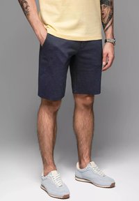 Pantaloni corti blu navy realizzati in un tessuto strutturato, con tasche e una vestibilità rilassata, abbinati a sneakers azzurro chiaro e una maglietta gialla.