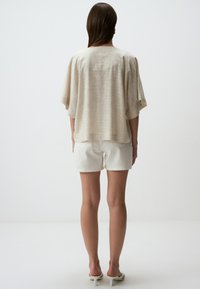 Blusa leggera beige a vestibilità morbida, maniche corte e tessuto strutturato; abbinata a pantaloni corti bianchi e sandali con punta aperta.