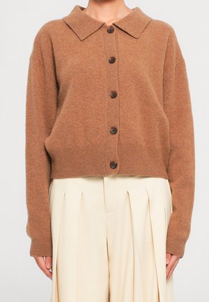 Cardigan marron boutonné à manches longues porté sur un pantalon crème plissé taille haute sur une personne devant un fond uni.
