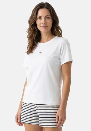 Vrouw met golvend bruin haar, die een wit T-shirt met korte mouwen draagt met een klein zwart logo en zwart-wit gestreepte korte broek, staand voor een effen achtergrond.