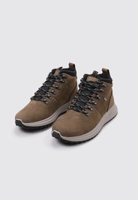 Par de botas de senderismo marrones con cordones blancos y negros, cuellos acolchados en el tobillo y suelas gruesas blancas sobre un fondo claro y sencillo.