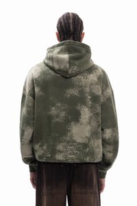 Olivově zelená mikina s kapucí s texturovaným abstraktním vzorem v různých odstínech zelené. Má prostornou kapuci a spuštěná ramena pro uvolněný střih.
