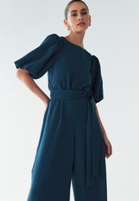  Een teal jumpsuit met een pofmouwen, een getailleerde bodice en een ceintuur om de taille. De stof is soepel met een zachte textuur en heeft een wijde pijpen ontwerp.