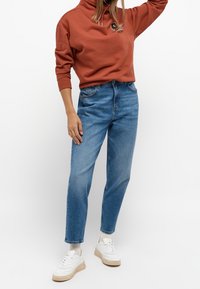 Sudadera de color óxido con un cuello alto, jeans de mezclilla azul de tiro alto y zapatillas deportivas blancas. La textura parece suave; la mezclilla tiene un acabado clásico.