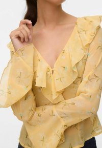 Blouse jaune transparente avec un motif floral, dotée d'un col et de manches froncés. Tissu léger avec une texture douce et fluide.