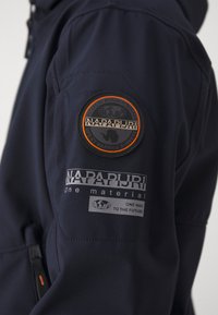 Rękaw granatowej kurtki z okrągłą naszywką Napapijri i napisami, w tym "One way to the future" oraz "One material".