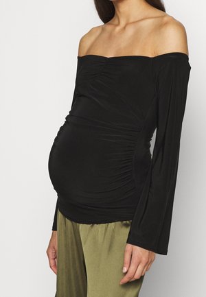 Zwangere persoon die een zwarte off-shoulder top met ruches en olijfgroene broeken draagt, staand tegen een effen witte achtergrond.
