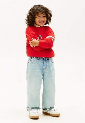 Uśmiechnięte dziecko o kręconych włosach, ubrane w czerwoną bluzę, jasnoniebieskie dżinsy i białe trampki, stojące z założonymi rękami na białym tle.