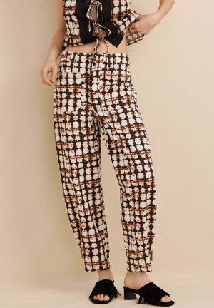 Vue recadrée d'une personne portant un pantalon à motif tie-dye avec des détails de bouton et de fermeture éclair, associé à des mules à talons ouvertes à bout ouvert avec franges noires.