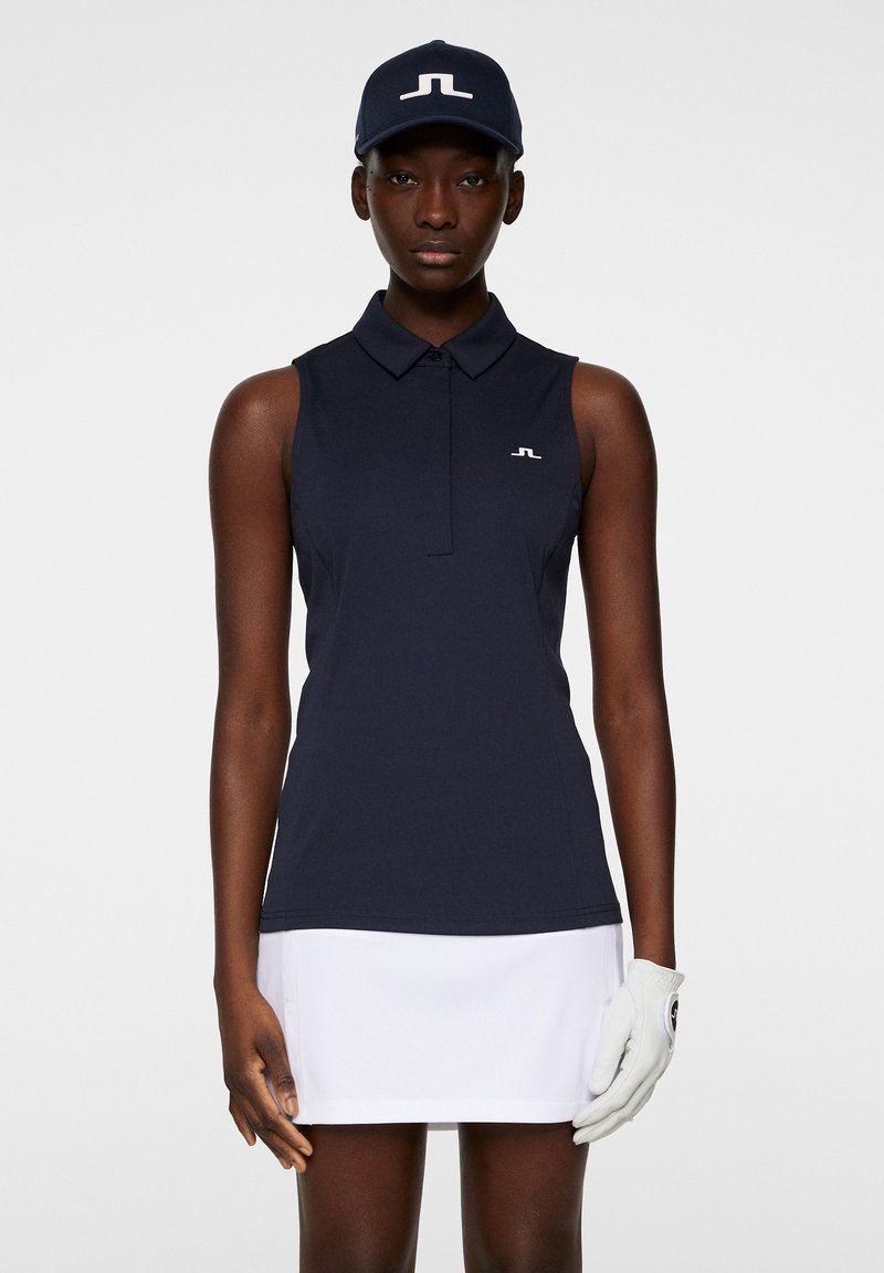 Marineblå ærmeløs polo-shirt med krave og hvidt logo. Kombineret med en hvid nederdel. Modellen har en matchende marineblå kasket og handske på.