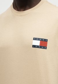 Κοντινή λήψη μπεζ t-shirt με ορθογώνιο λογότυπο Tommy Jeans σε λευκό, κόκκινο και μαύρο στο πάνω μέρος του στήθους.