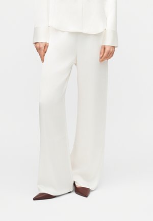 Žena oblečená v voľných bielych nohaviciach, bielom dlhom rukáve a hnedých špicatých topánkach, stojaca pred jednoduchým svetlým pozadím.