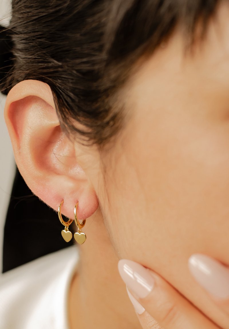 Pendientes de aro dorados con pequeños charms en forma de corazón, con un acabado brillante y un diseño sencillo, llevados en el lóbulo cerca de la oreja.