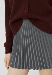 Cardigan en maille bourgogne sur une jupe plissée grise. La jupe présente des plis verticaux et un tissu texturé avec une finition brillante.