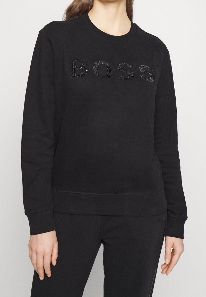 Personne portant un sweat-shirt noir à manches longues avec le mot "BOSS" écrit en petits clous noirs sur la poitrine.