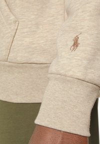 Světle béžová mikina s texturovaným materiálem, žebrovanými manžetami a malým hnědým logem polokošile na rukávu. Žádné viditelné vzory ani akcenty.