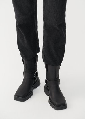 Black leather ankle boots with a square toe, featuring a wide strap and metal buckle detail. The boots have a chunky sole and smooth texture.