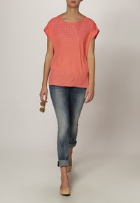 Koralfarvet kortærmet top med tekstureret detalje omkring udskæringen, kombineret med lyseblå opsmøgede jeans og nude ballerinaer. Solbriller holdt i hånden.