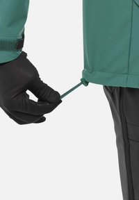 Hand in black glove tightening drawstring on green jacket hem, showing adjustable outdoor clothing detail against white background.