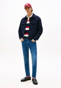 Noor mees punase nokamütsi, tumesinise jaki, triibulise polo-särgi, siniste teksade, punaste sokkide ja mustade paeltega kingadega, seisab käed taskutes.