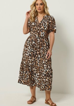 Kvinde iført en brun og sort leopardprint midi-kjole med korte ærmer og lyse flade sandaler, stående mod en ensfarvet lys baggrund.