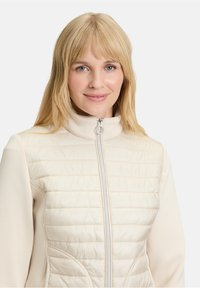 Kremowa puchowa kurtka z wysokim kołnierzem wyłożonym sztucznym futrem, z zamkiem błyskawicznym oraz fakturowanym pikowanym przodem, kontrastującym z gładkimi rękawami.
