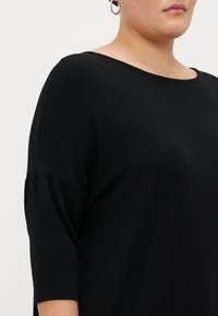 Crni top od mekog materijala, s wide izrezom, kratkim rukavima i opuštenim krojem, s glatkom teksturom i bez vidljivih uzoraka.