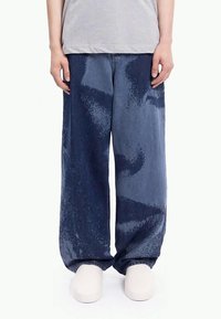 Άτομο που φοράει φαρδιά μπλε τζιν με tie-dye, ανοιχτό γκρι πουκάμισο και λευκά παντόφλες, στέκεται μπροστά σε λευκό φόντο.