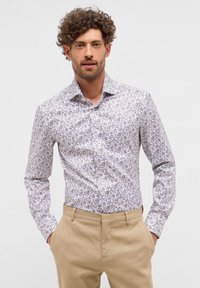 Weißes Hemd mit floralem Muster in sanften lila Tönen. Langärmlig, mit Knopfleiste, getragen mit khakifarbener Hose. Sanfte Stofftextur.