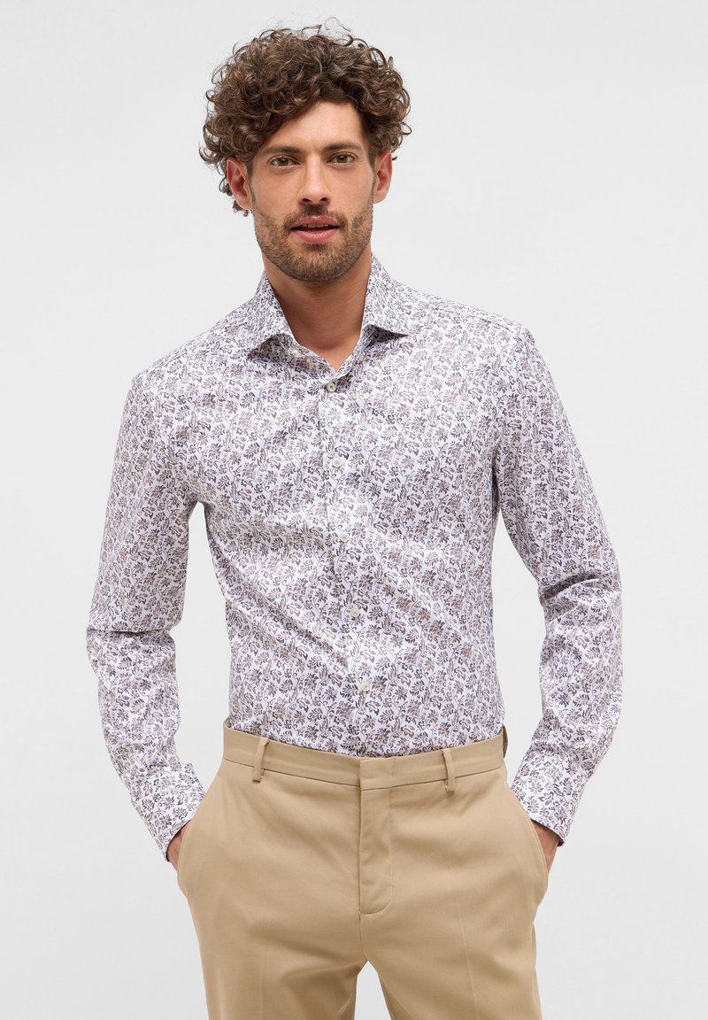 Weißes Hemd mit floralem Muster in sanften lila Tönen. Langärmlig, mit Knopfleiste, getragen mit khakifarbener Hose. Sanfte Stofftextur.