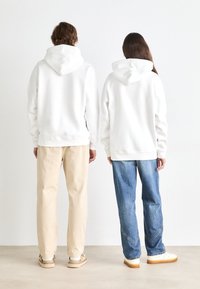Two people wear white hoodies, standing back-to-back. The hoodies have front pockets and are paired with beige and blue jeans, respectively.