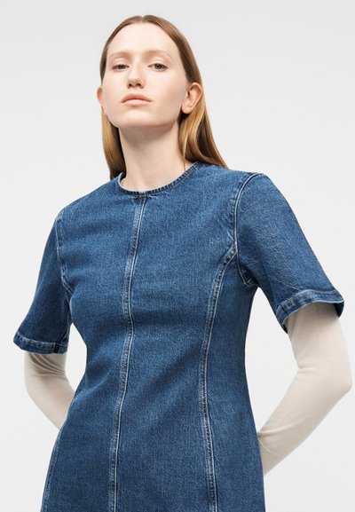 Γυναίκα με ανοιχτόχρωμο δέρμα και ίσια ανοιχτό καστανά μαλλιά φοράει εφαρμοστό κοντομάνικο φόρεμα από τζιν, πάνω από μακρυμάνικο μπεζ πουκάμισο, πάνω σε απλό φόντο.