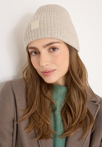 Gorro de punto beige con textura acanalada y un parche con logo, llevado por una persona con cabello largo castaño, combinado con un suéter verde y un abrigo marrón.