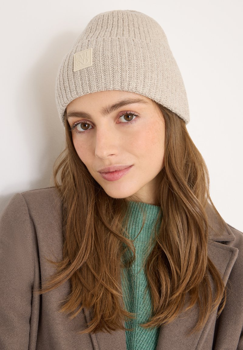 Gorro de punto beige con textura acanalada y un parche con logo, llevado por una persona con cabello largo castaño, combinado con un suéter verde y un abrigo marrón.