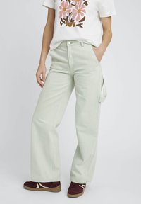 Pantalons à larges jambes vert clair en denim, dotés de poches latérales et d'une coupe décontractée. Associés à un t-shirt graphique blanc et des baskets marron.