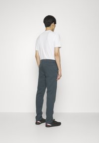Pantalones oscuros en teal para hombre, confeccionados en una mezcla de algodón, con un corte ajustado, un bolsillo trasero y un logo sutil. El modelo lleva una camiseta blanca lisa.