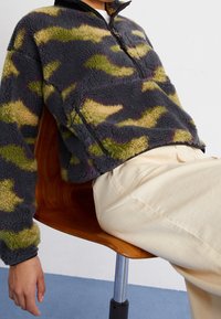 Polarowy pulower w ciemnoszarym kolorze z kamuflażowym wzorem w zieleni i beżu. Posiada wysoki kołnierz, zamek błyskawiczny oraz boczne kieszenie.