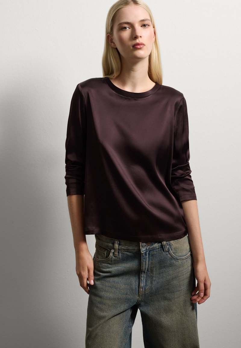 Giovane donna con lunghi capelli biondi che indossa un top a maniche lunghe di un viola scuro lucido e jeans blu sbiaditi a vestibilità ampia contro un muro semplice.