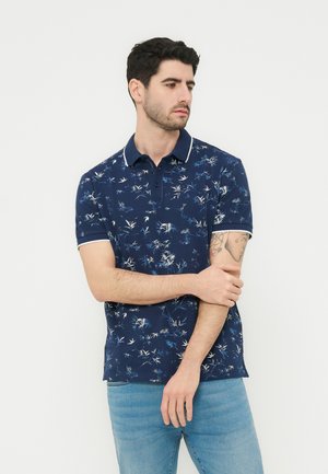 Άνδρας με σκούρα μαλλιά που φορά μπλε ναυτικό πόλο με φλοράλ σχέδιο και ανοιχτό μπλε τζιν, κρατώντας τον αριστερό του βραχίονα και κοιτάζοντας προς τα πλάγια.