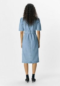 Vestido de mezclilla en azul claro, hasta la rodilla, con mangas cortas abullonadas, cintura ceñida por un cinturón y corte recto. Presenta costuras reforzadas.