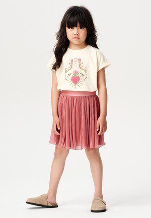 Camicia crema a maniche corte con grafica a cuore floreale, abbinata a una gonna rosa plissettata. Il modello indossa delle scarpe beige senza lacci.