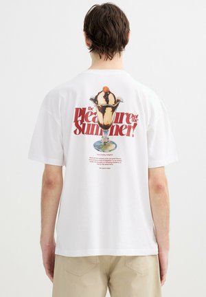 Hombre con camiseta blanca con gráfico de helado sundae y texto rojo "¡El placer del verano!" en la espalda.