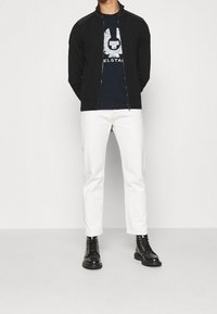 Schwarze Zip-Jacke mit hohem Kragen, navyfarbiges Grafik-T-Shirt, weiße Slim-Fit-Hosen und schwarze Schnürstiefel. Glattes Material und taillierte Passform.