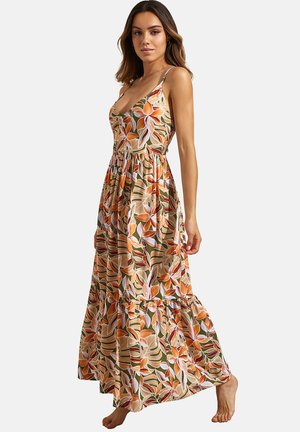 Femme portant une robe maxi sans manches à motifs floraux orange, verts et blancs, debout pieds nus devant un fond uni.