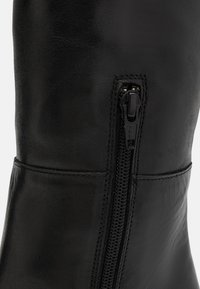 Zbliżenie czarnego skórzanego buta z pionowym czarnym zamkiem błyskawicznym i widocznymi szwami wzdłuż górnej i bocznej krawędzi.