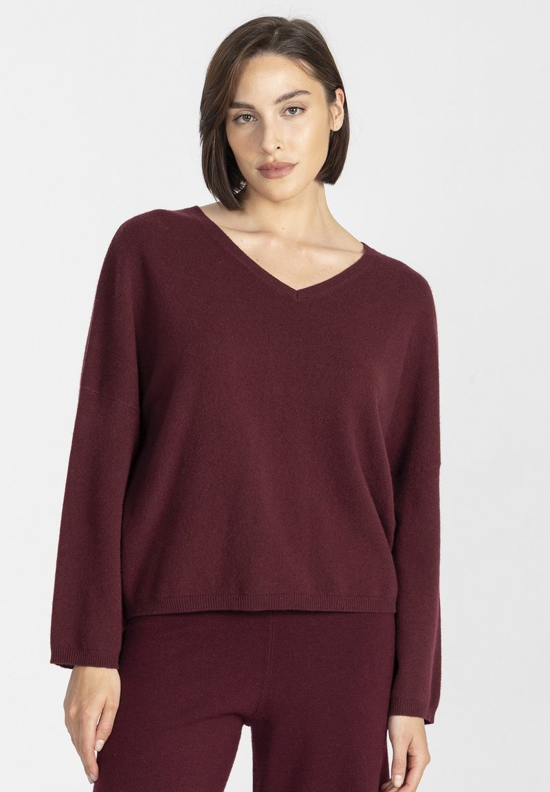 Bordowy sweter w serek z miękkiego materiału, o luźnym kroju, z długimi rękawami i żeberkowym wykończeniem. W zestawie z pasującymi spodniami.