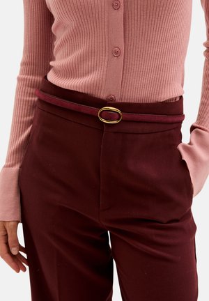 Mujer vistiendo una blusa de botones de ribete rosa metida en unos pantalones de tiro alto color marrón con un cinturón delgado a juego y una hebilla ovalada dorada.