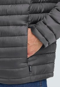Grå pufferjakke med quiltet design, som har en sidelomme og glidelås. Laget av glatt, lett materiale.